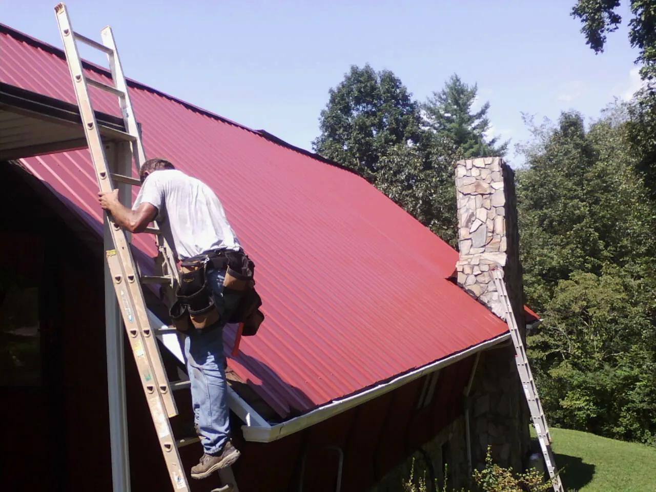 Roofer on a ladder beside a red metal roof and stone chimney — Soffit & Fascia Repair by Roofing Brothers in Strawberry Point